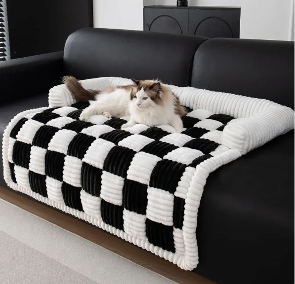 A fluffy cat lounges on a cozy, black and white checkered CozyPlush Pet Bed with a plush white border, set on a black sofa.